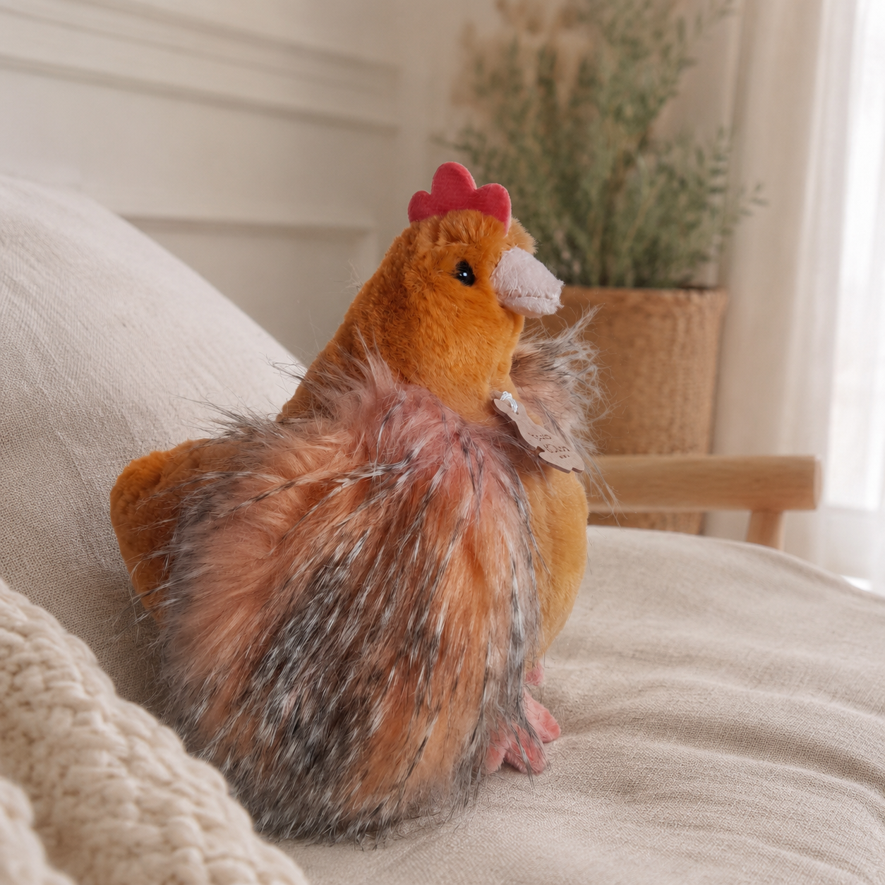 A plush chicken toy with orange-brown feathers, a pink comb, and gray bristles, sitting on a beige fabric surface in a softly lit indoor setting.
